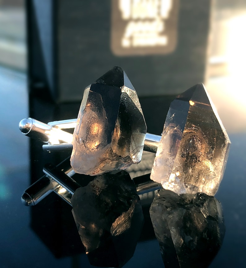 May include: A pair of silver cufflinks with clear, faceted crystal accents. The crystals are shaped like points and have a smoky gray color.