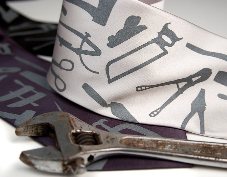May include: A white silk tie with a gray pattern of various tools, including a wrench, pliers, and a saw. The tie is lying on top of a purple fabric with a wrench on top of it.