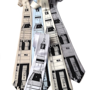 May include: Four silk ties featuring a black and white illustration of a rocket ship with the text "USA" printed on each tie. The ties are in different colors: beige, light blue, white, and gray.