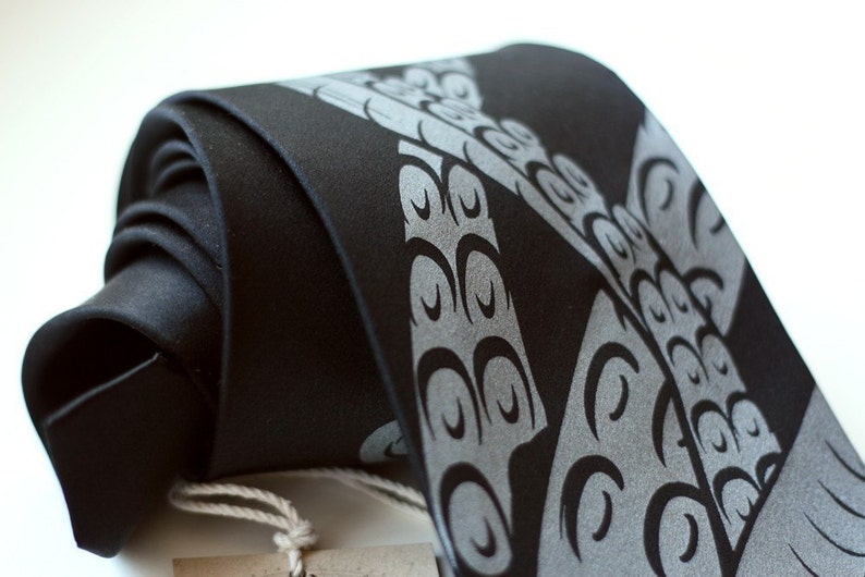 Puede incluir: Una corbata negra con un patr&oacute;n de ondas estilizadas de color gris plateado. La corbata est&aacute; doblada y anudada.
