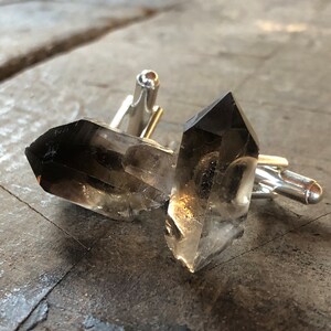 May include: A pair of cufflinks featuring two clear quartz crystals with a smoky grey hue. The crystals are attached to silver-toned metal cufflink backs.