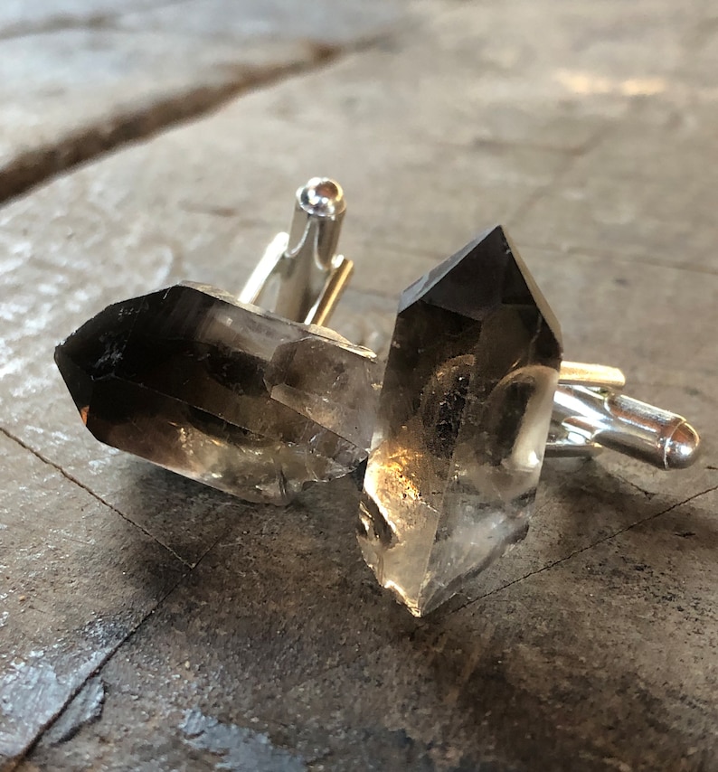 May include: A pair of cufflinks featuring two clear quartz crystals with a smoky grey hue. The crystals are attached to silver-toned metal cufflink backs.