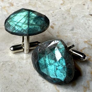 May include: A pair of cufflinks featuring faceted labradorite gemstones in shades of teal and gray. The stones are mounted on silver-colored metal posts with a rounded toggle closure. The gemstones have a unique, iridescent quality.