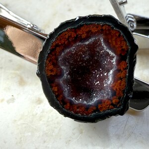 May include: A tie clip featuring a circular geode slice with a dark black outer edge, a vibrant orange-red ring, and a sparkling, crystalline center. The geode is securely mounted onto a polished, silver-toned metal tie bar with a spring-loaded clasp.