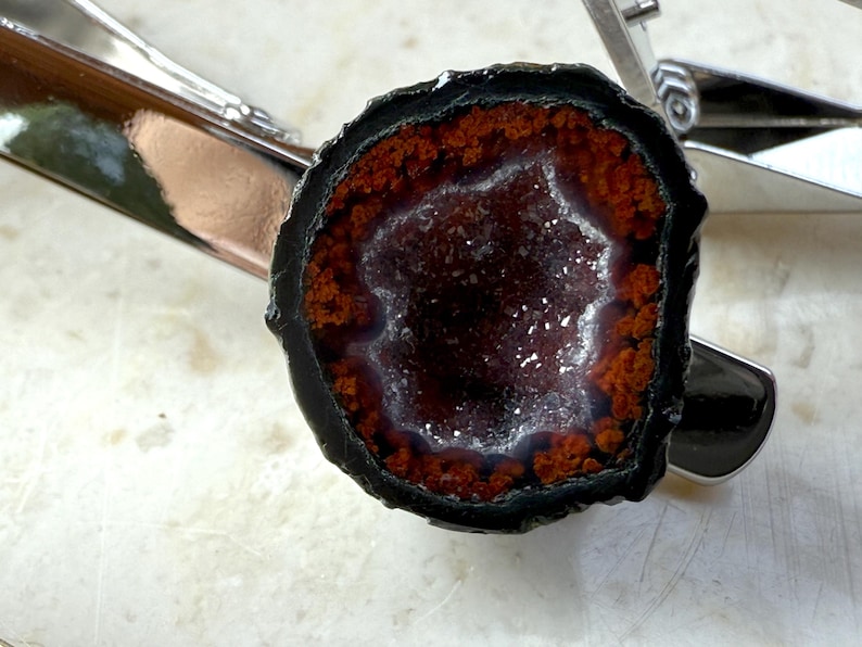 May include: A tie clip featuring a circular geode slice with a dark black outer edge, a vibrant orange-red ring, and a sparkling, crystalline center. The geode is securely mounted onto a polished, silver-toned metal tie bar with a spring-loaded clasp.