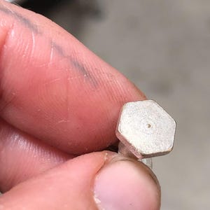 May include: A small, silver-colored, hexagonal-shaped bolt with a central hole. The bolt is held between fingers, with the focus on the bolt's shape and metallic finish. The background is blurred, emphasizing the bolt's details.
