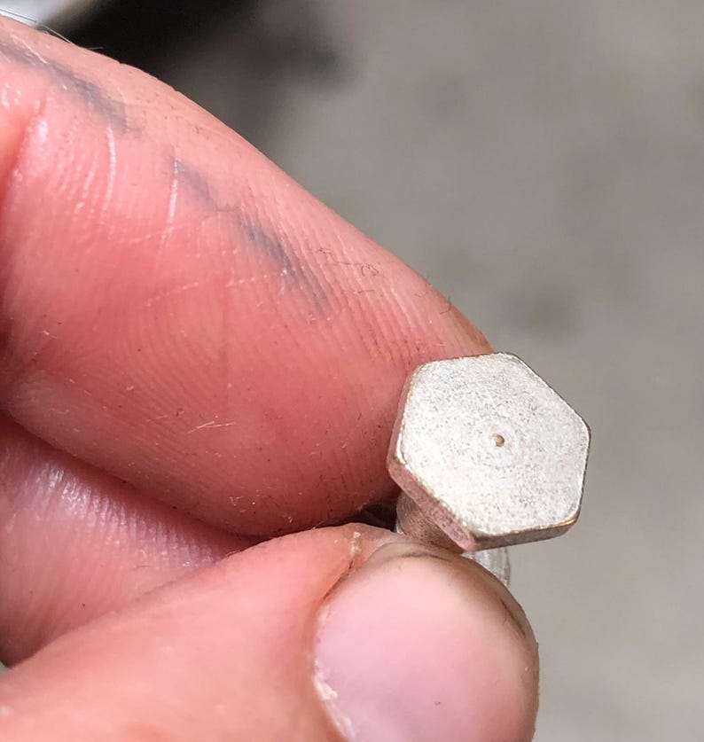 May include: A small, silver-colored, hexagonal-shaped bolt with a central hole. The bolt is held between fingers, with the focus on the bolt's shape and metallic finish. The background is blurred, emphasizing the bolt's details.
