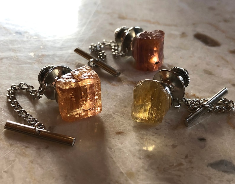 May include: Three tie tacks with different colored gemstones. The gemstones are all cut into different shapes. The tie tacks are made of silver metal.