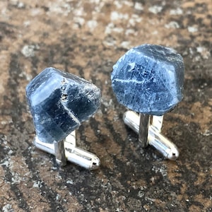 May include: A pair of cufflinks featuring raw blue sapphire gemstones set in silver.
