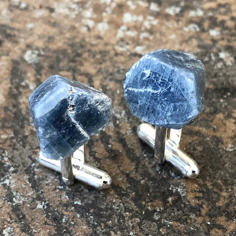 May include: A pair of cufflinks featuring raw blue sapphire gemstones set in silver.