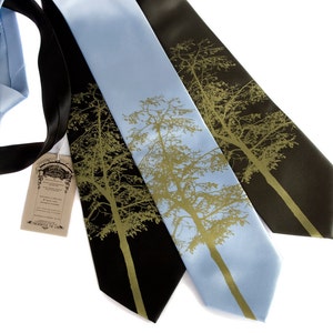 May include: Three neckties in black, light blue, and dark brown, each featuring a gold tree design. A light blue tie is partially visible on the left. A tag with text hangs from the black tie. The ties are displayed against a white background.