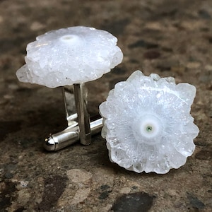 May include: A pair of cufflinks featuring white crystal formations with a silver metal base. The crystals have a unique, flower-like shape.