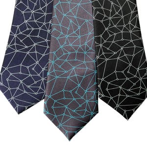 May include: Three neckties with geometric patterns. One is navy blue with a white design, another is gray with a teal design, and the third is black with a white design. The ties are arranged on a white background.