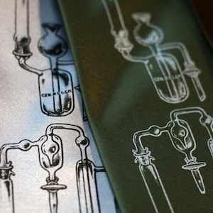 May include: Two silk ties featuring a white scientific illustration of a distillation apparatus. The ties are green and silver.