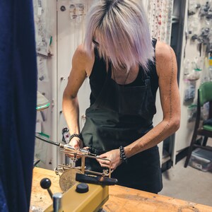 May include: A person working in a workshop, wearing a black apron, using tools on a metal object. The workshop has a wooden table, a drill press, and various tools and supplies. The person has light purple hair.