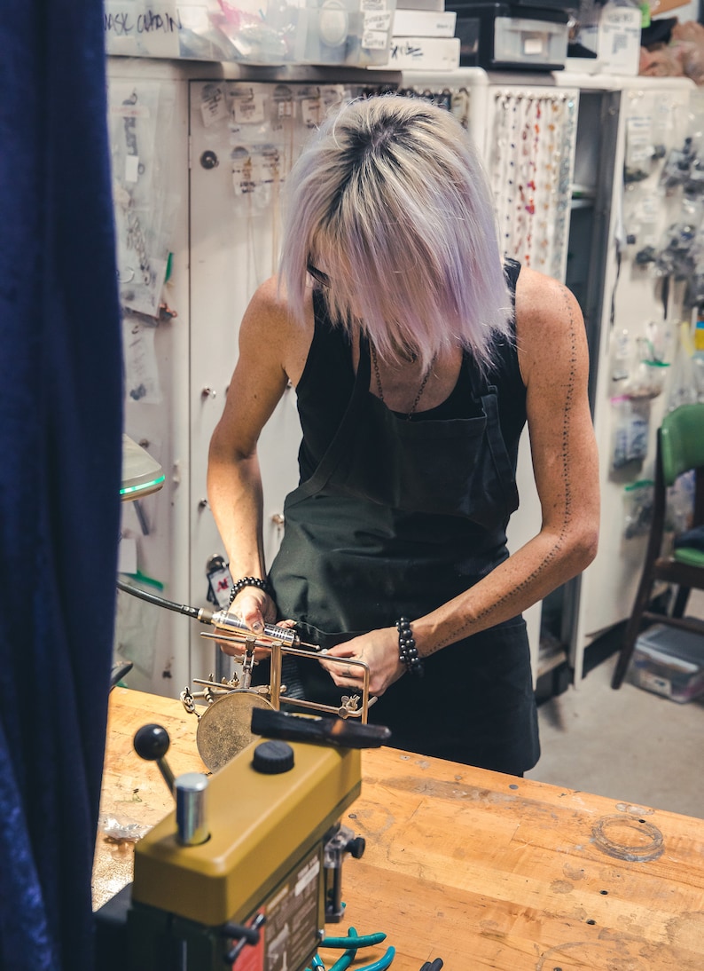 May include: A person working in a workshop, wearing a black apron, using tools on a metal object. The workshop has a wooden table, a drill press, and various tools and supplies. The person has light purple hair.