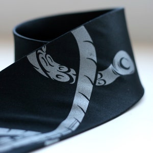 Puede incluir: Diadema de tela negra con un dise&ntilde;o plateado estilizado de una planta con un tallo curvo y hojas.