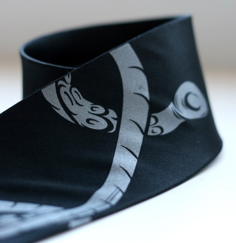 Puede incluir: Diadema de tela negra con un dise&ntilde;o plateado estilizado de una planta con un tallo curvo y hojas.