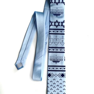 May include: A light blue tie with a repeating pattern of navy blue Stars of David, menorahs, dreidels, and hearts. The tie is made of a smooth, satin-like material and is displayed on a white background.