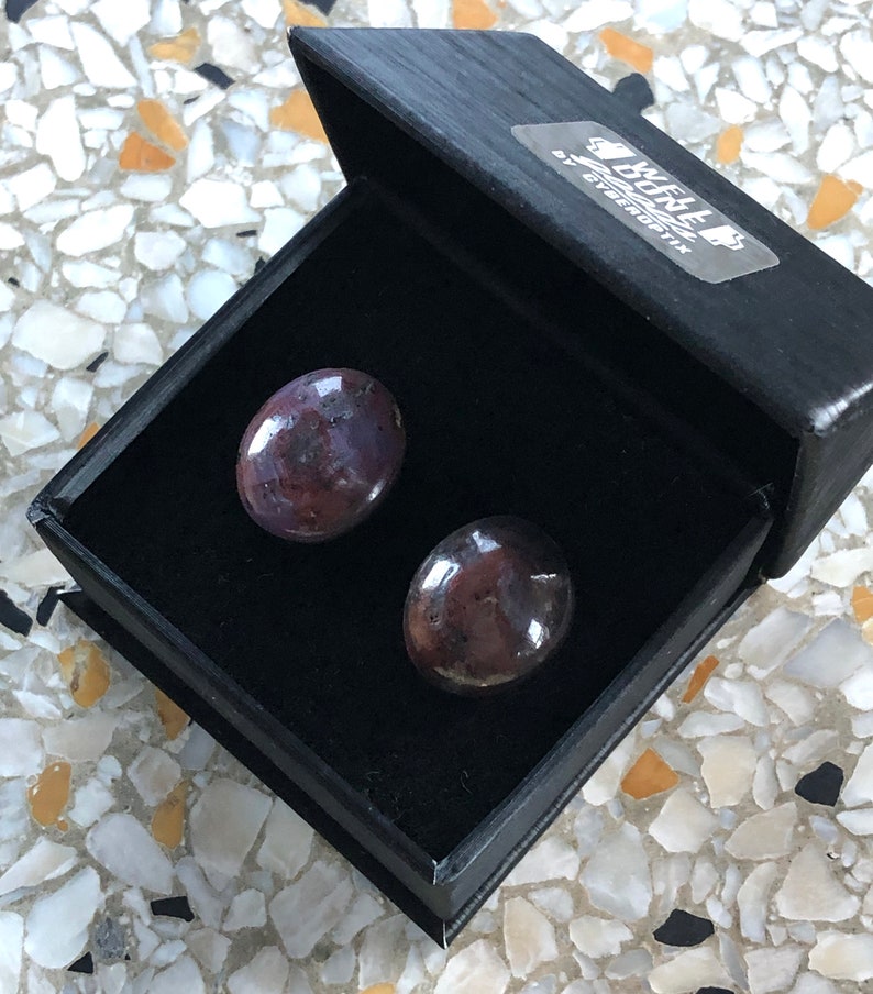 K&ouml;nnte beinhalten: Zwei runde Ohrringe aus dunkelviolettem und schwarzem Edelstein in einer schwarzen, mit Samt ausgekleideten Schachtel. Die Schachtel ist mit "We Love Buttons" beschriftet, einem kleinen Herz und dem Text "By Everginix".