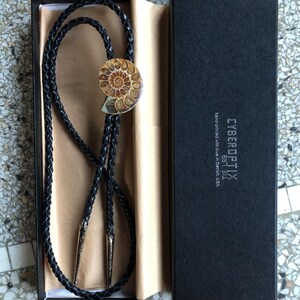 May include: A black braided leather bolo tie featuring a polished spiral ammonite fossil centrepiece and silver-toned metal tips. The tie is presented in a black rectangular box labelled CYBEROPTIX TIE LAB hand-printed with love in Detroit, USA.