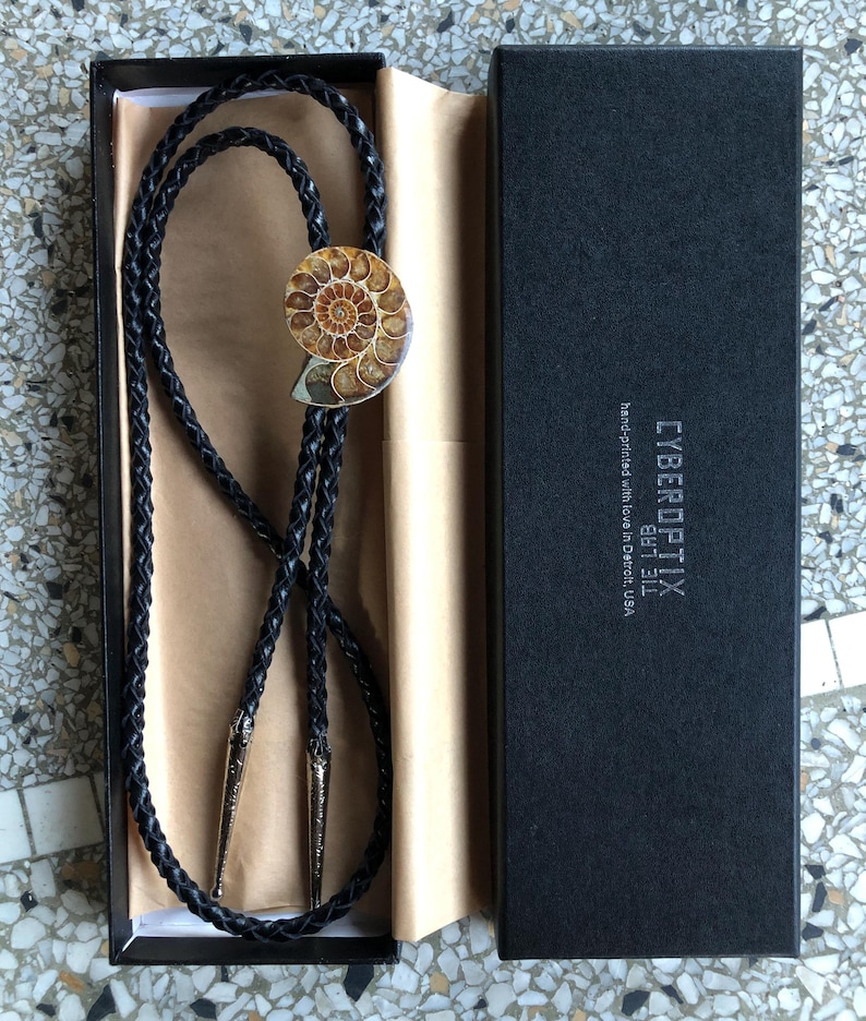 May include: A black braided leather bolo tie featuring a polished spiral ammonite fossil centrepiece and silver-toned metal tips. The tie is presented in a black rectangular box labelled CYBEROPTIX TIE LAB hand-printed with love in Detroit, USA.