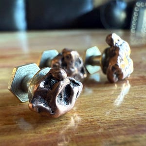 May include: Close-up of four copper-colored, irregularly shaped cabinet knobs with brass hardware. The knobs have a textured, organic appearance, suggesting a natural or handcrafted design. They are set against a blurred wooden surface.