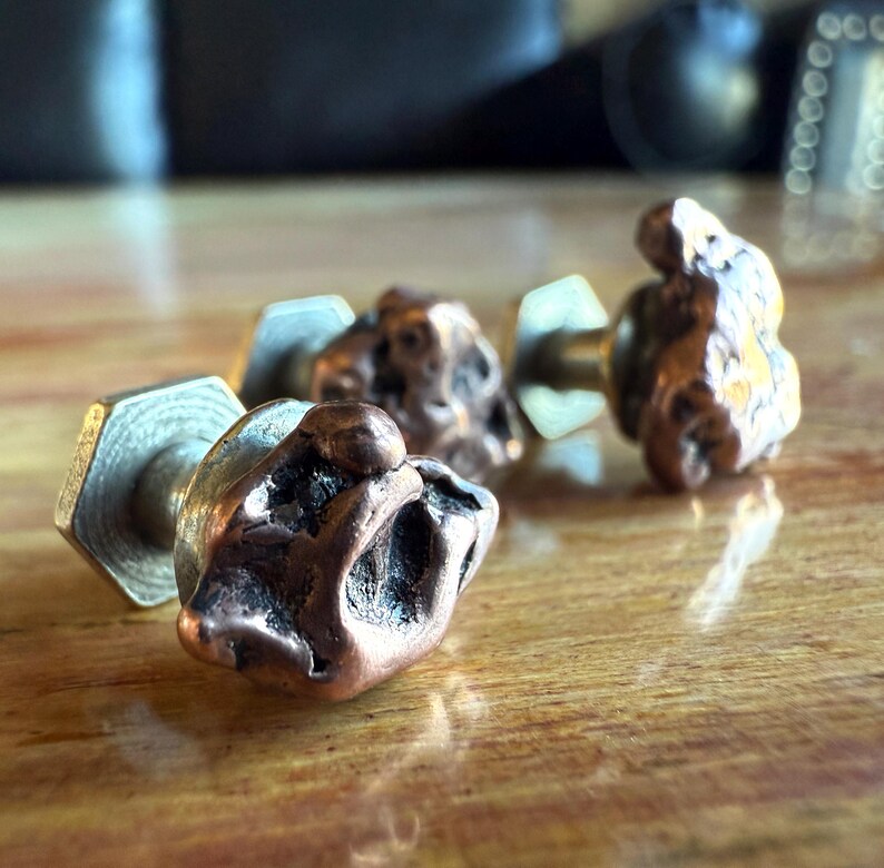 May include: Close-up of four copper-colored, irregularly shaped cabinet knobs with brass hardware. The knobs have a textured, organic appearance, suggesting a natural or handcrafted design. They are set against a blurred wooden surface.