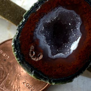 May include: A tie clip featuring a circular geode slice with a dark green outer edge, a deep reddish-brown band, and a central cavity filled with sparkling white crystals. A copper-colored 2022 penny is positioned next to the geode for scale.