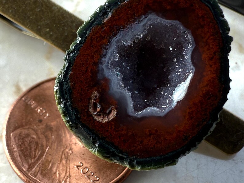 May include: A tie clip featuring a circular geode slice with a dark green outer edge, a deep reddish-brown band, and a central cavity filled with sparkling white crystals. A copper-colored 2022 penny is positioned next to the geode for scale.