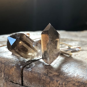 May include: A pair of cufflinks featuring two clear, smoky quartz crystals. The crystals are faceted and have a pointed shape. The cufflinks are made of silver metal.