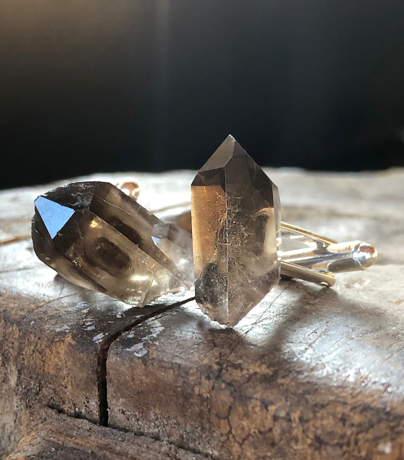 May include: A pair of cufflinks featuring two clear, smoky quartz crystals. The crystals are faceted and have a pointed shape. The cufflinks are made of silver metal.