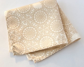 May include: A beige pocket square with a white lace pattern. The fabric is folded in half and the edges are stitched.