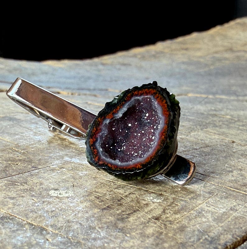 May include: A silver-toned metal tie clip featuring a circular geode slice. The geode has a dark, crystalline purple center surrounded by a thin white ring and a vibrant orange-red border, encased in a dark, textured, natural outer edge.
