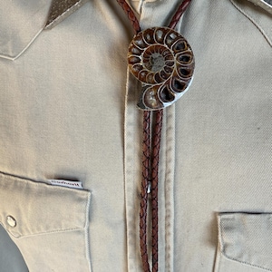 May include: A bolo tie featuring a polished brown ammonite fossil centrepiece mounted on a braided dark brown leather cord. The cord is secured with silver-toned metal tips and is worn over a tan button-down shirt with a Carhartt brand label.