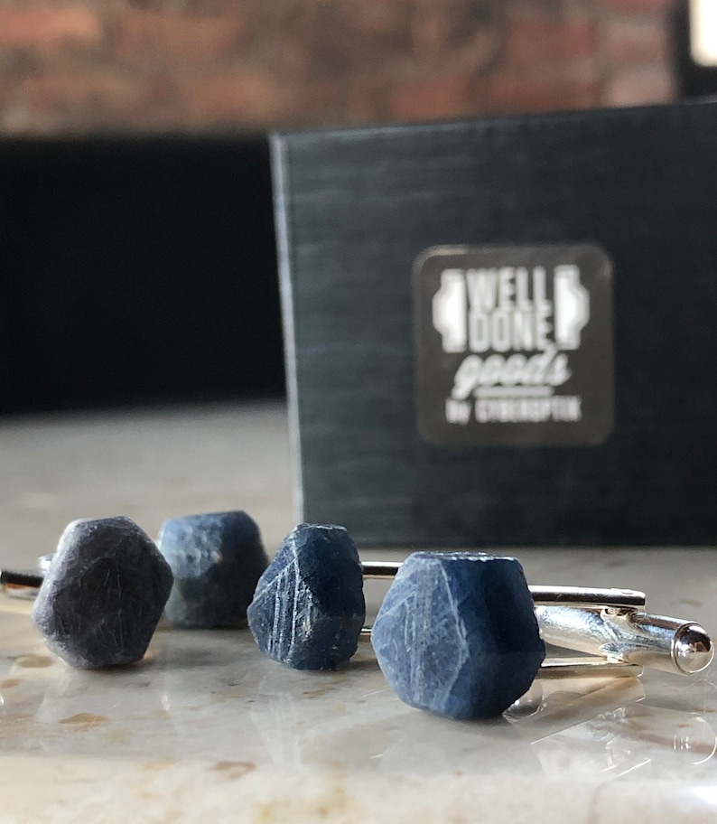 May include: A set of four cufflinks featuring blue gemstone accents. The cufflinks are silver and have a polished finish. The gemstones are faceted and have a dark blue color. The cufflinks are displayed on a white surface. The text "WELL DONE goods by CYBERKRAFTIK" is visible on a black box in the background.