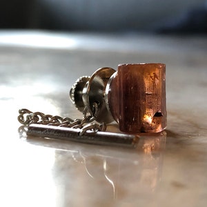 May include: A silver tie tack with a chain and a clear, rectangular, pink-hued stone.