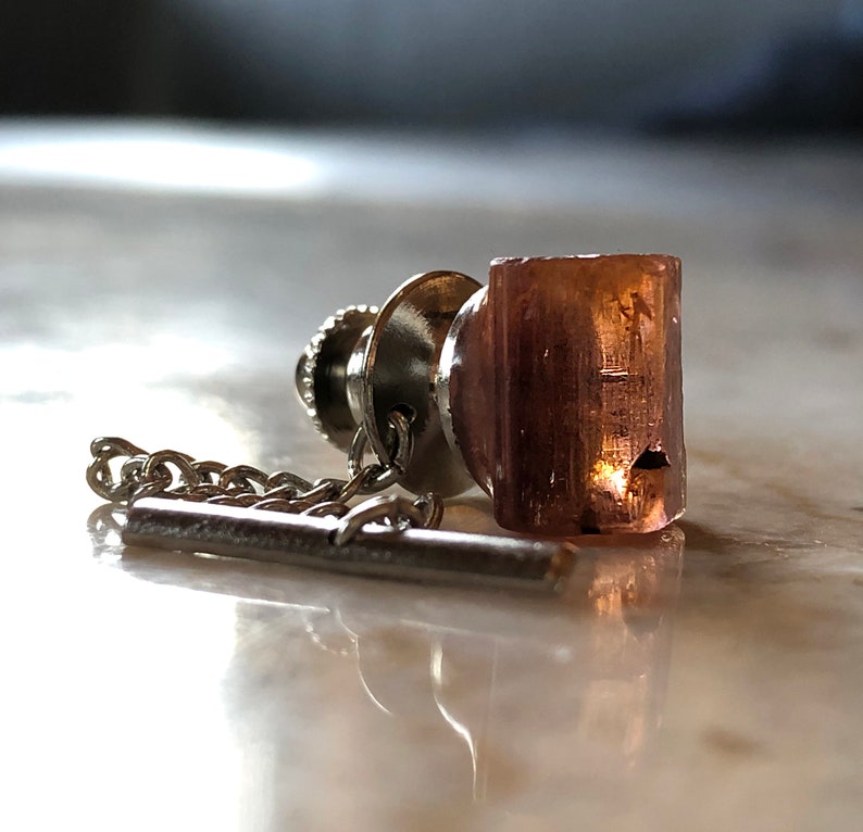 May include: A silver tie tack with a chain and a clear, rectangular, pink-hued stone.
