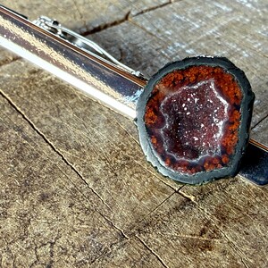 May include: A silver-toned metal tie clip featuring a circular geode slice. The geode has a dark black outer edge, a deep reddish-brown ring, and a sparkling, crystalline center with small white and purple mineral deposits throughout the cavity.