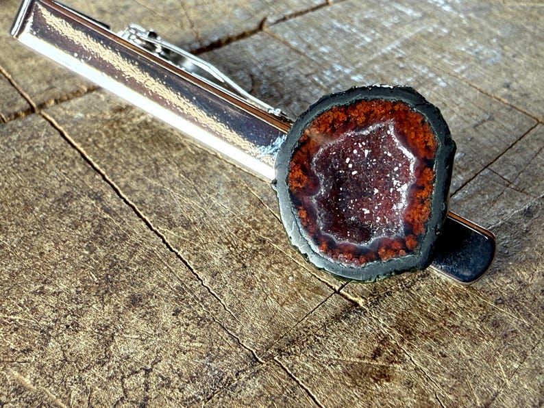 May include: A silver-toned metal tie clip featuring a circular geode slice. The geode has a dark black outer edge, a deep reddish-brown ring, and a sparkling, crystalline center with small white and purple mineral deposits throughout the cavity.