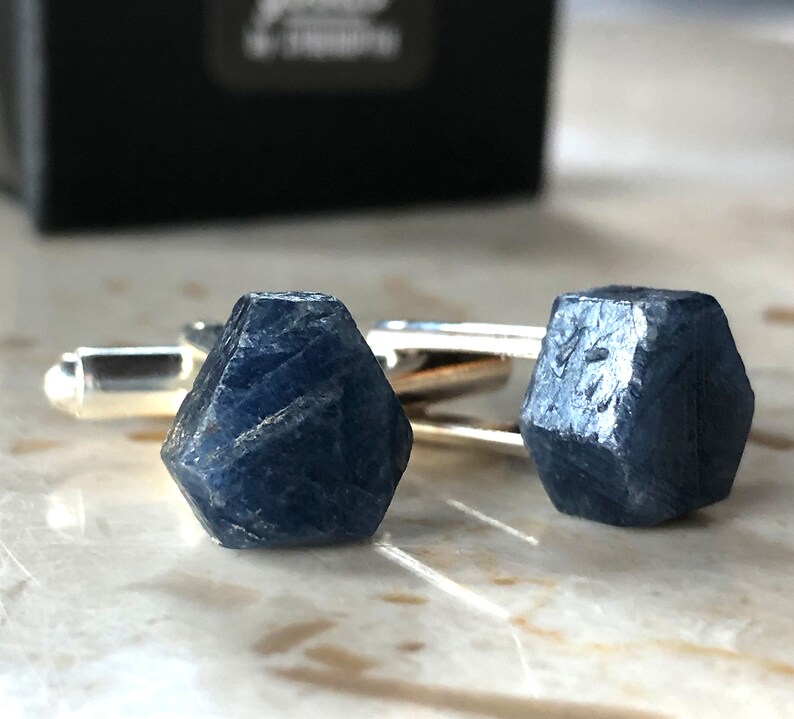 May include: A pair of cufflinks featuring a dark blue sapphire gemstone in a faceted, geometric shape. The cufflinks are made of silver.