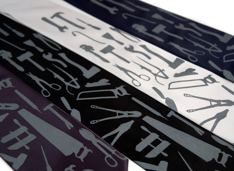 May include: Four neckties with a repeating pattern of tools in a silver color on different colored backgrounds. The ties are navy blue, white, black, and purple.
