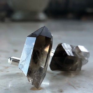 May include: A pair of cufflinks made from smoky quartz crystals. The crystals are faceted and have a dark gray color. The cufflinks are attached to silver metal.