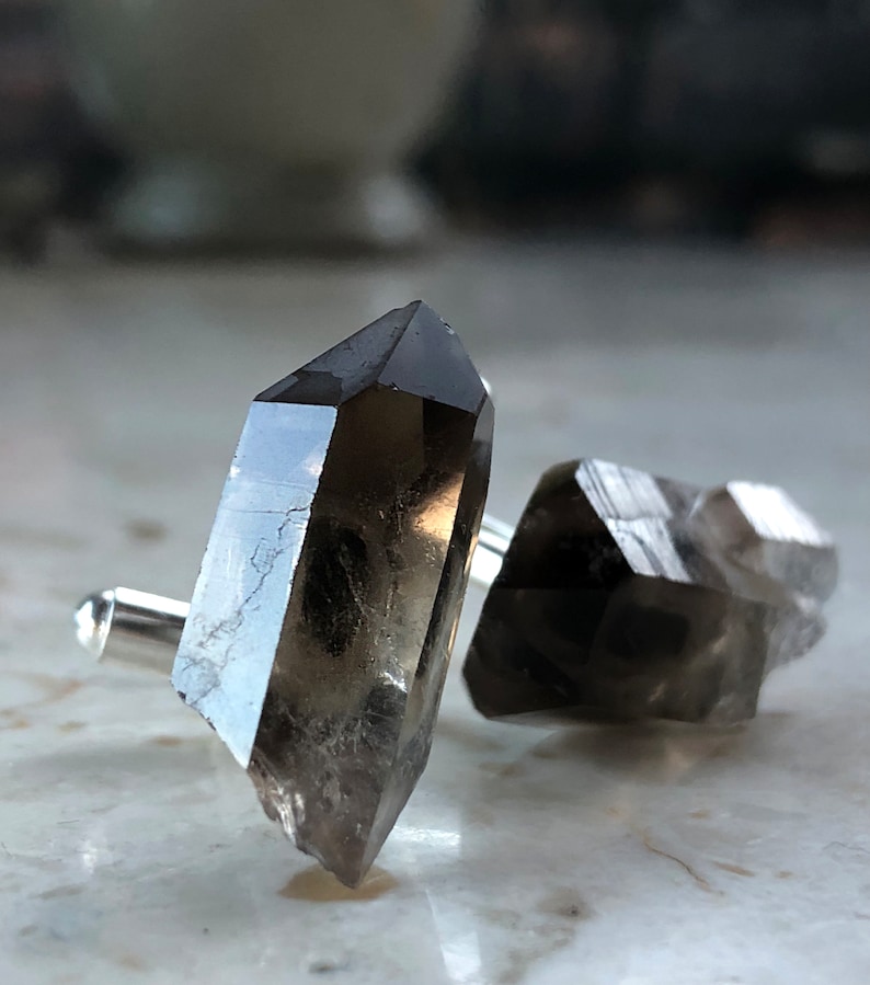 May include: A pair of cufflinks made from smoky quartz crystals. The crystals are faceted and have a dark gray color. The cufflinks are attached to silver metal.