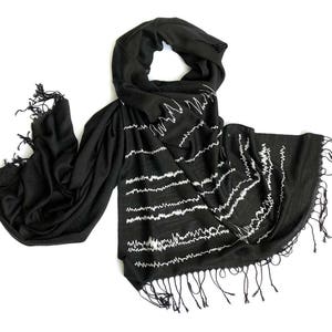 Puede incluir: Bufanda negra con patrones bordados blancos en forma de onda. La bufanda tiene forma rectangular con bordes con flecos. El diseño presenta líneas horizontales con un patrón ondulado, creando una textura visual única. La bufanda está hecha de un material suave y fluido.