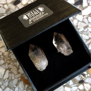 May include: Two clear quartz crystal points in a black box with a label that reads "WELL DONE goods by CYBEROPTIX".