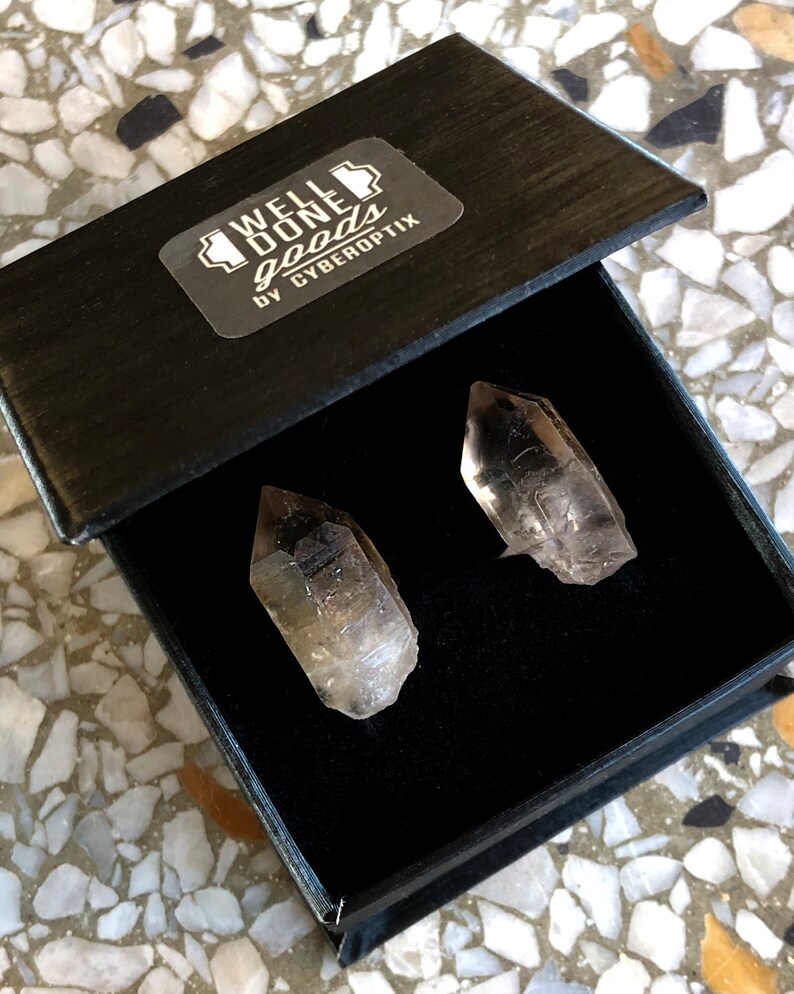 May include: Two clear quartz crystal points in a black box with a label that reads "WELL DONE goods by CYBEROPTIX".