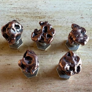 May include: Five copper-colored, irregularly shaped cabinet knobs. Each knob is mounted on a silver-colored, hexagonal base. The knobs have a textured, organic appearance, suggesting a natural or handcrafted design.