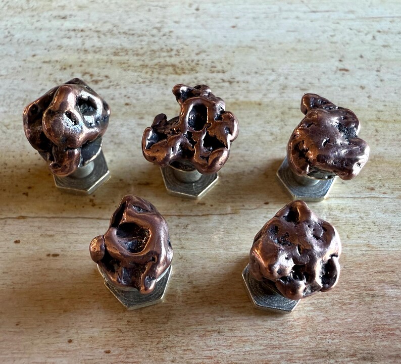 May include: Five copper-colored, irregularly shaped cabinet knobs. Each knob is mounted on a silver-colored, hexagonal base. The knobs have a textured, organic appearance, suggesting a natural or handcrafted design.