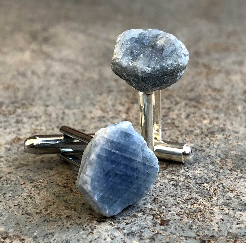 May include: A pair of cufflinks featuring raw blue sapphire gemstones. The sapphires are set in silver metal.